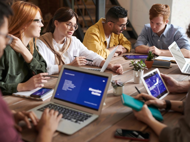 a group of people sitting around a table with laptops and tablets